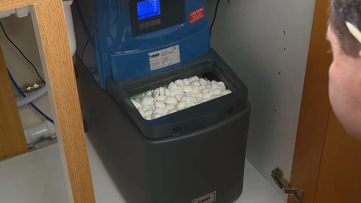 Blue water softener in a cabinet with its lid off, showing white salt pellets, and a man looking in